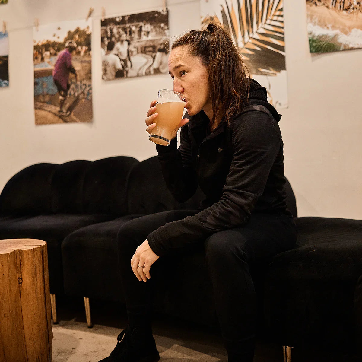 Woman in GORUCK grid fleece hoodie drinking juice, sitting on black couch indoors