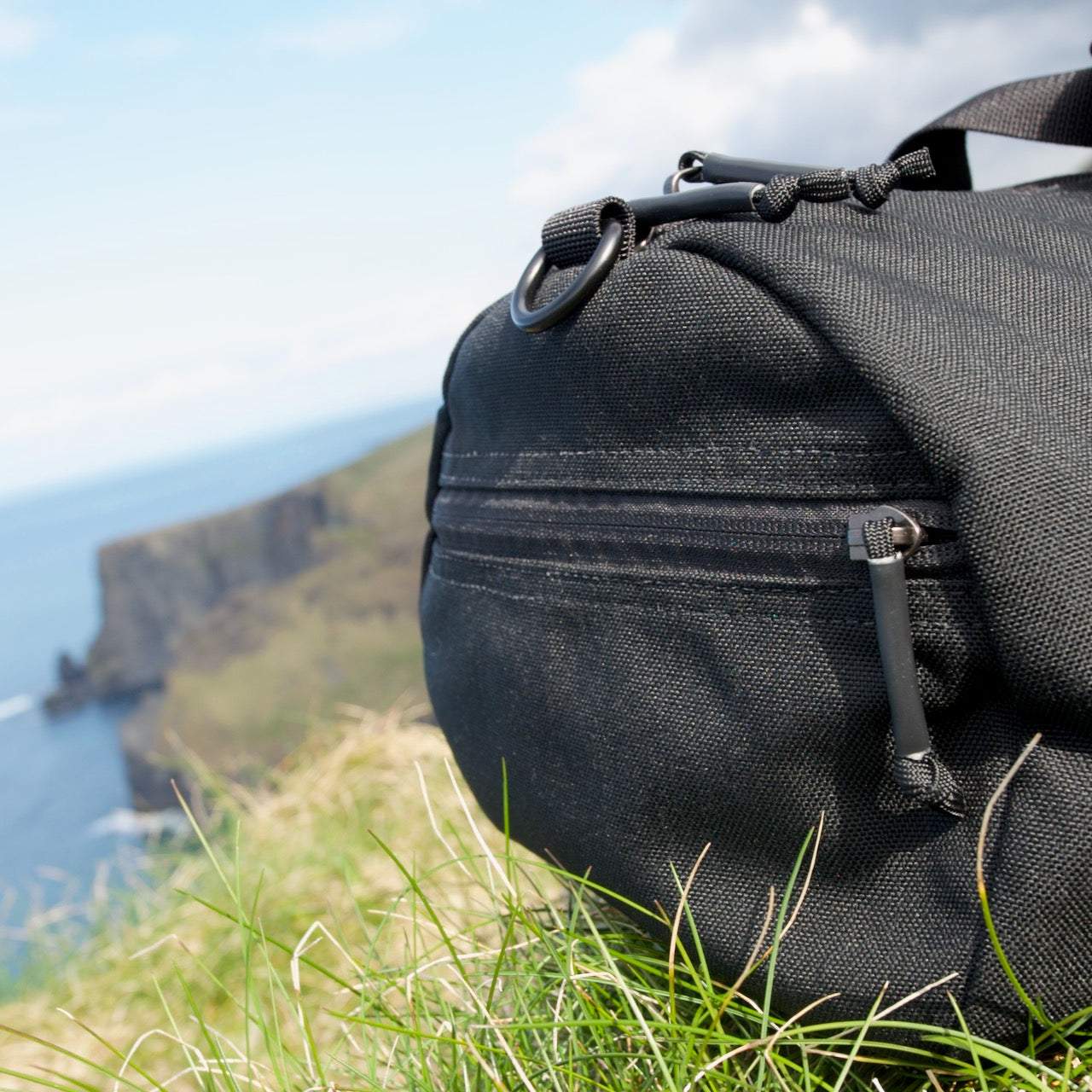Close-up of the Gym Bag - Cordura in black, crafted from durable 1000D CORDURA, resting on grassy ground with cliffs and the ocean softly blurred in the background.