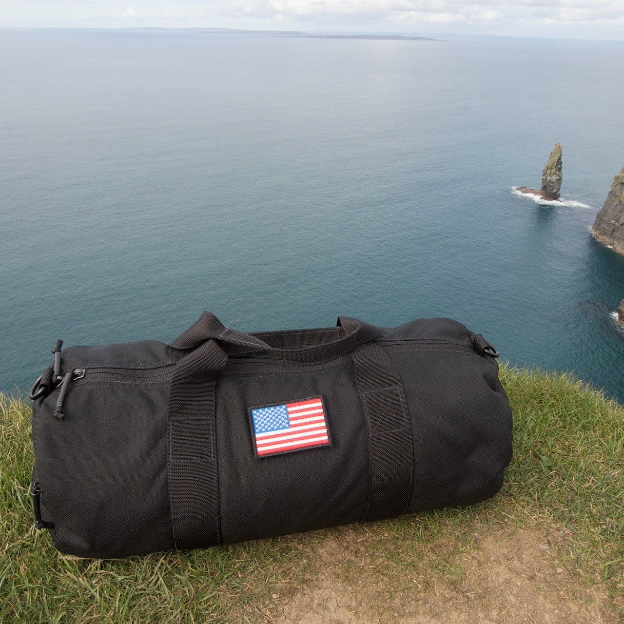 A black Gym Bag - Cordura with a US flag patch rests on a grassy cliff by the ocean and rock formations—a rugged, adventure-ready duffel for organizing gym gear.
