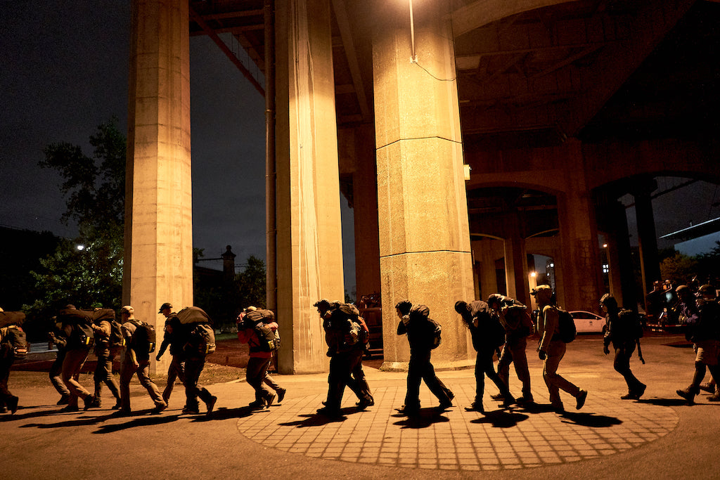 A line of people walk under a highway overpass at night, illuminated by yellow streetlights.