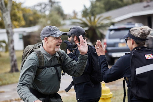 Two people wearing backpacks high-five each other outdoors near a house and a parked SUV.