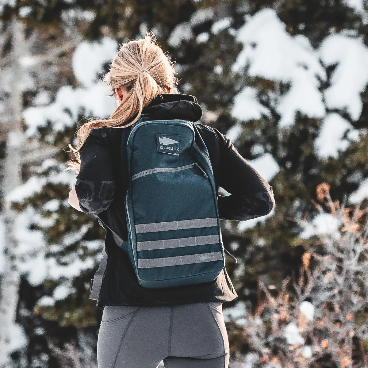 Woman outdoors wearing dark sea blue GORUCK backpack in snowy forest setting