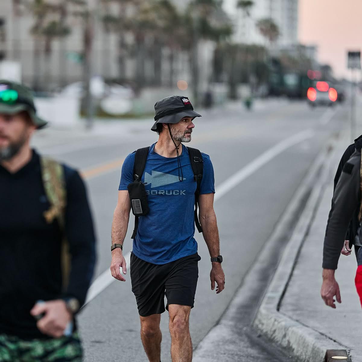 Man walking on street wearing charcoal GORUCK boonie hat, blue GORUCK t-shirt, and black shorts with backpack