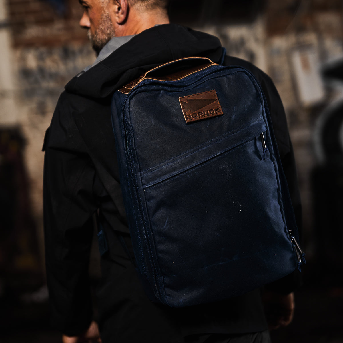 A person stands in an urban setting, wearing a black jacket and carrying a Heritage Rucksack, the navy GR1 USA - Heritage backpack crafted from Waxed Duck Canvas with brown detailing for enduring style.