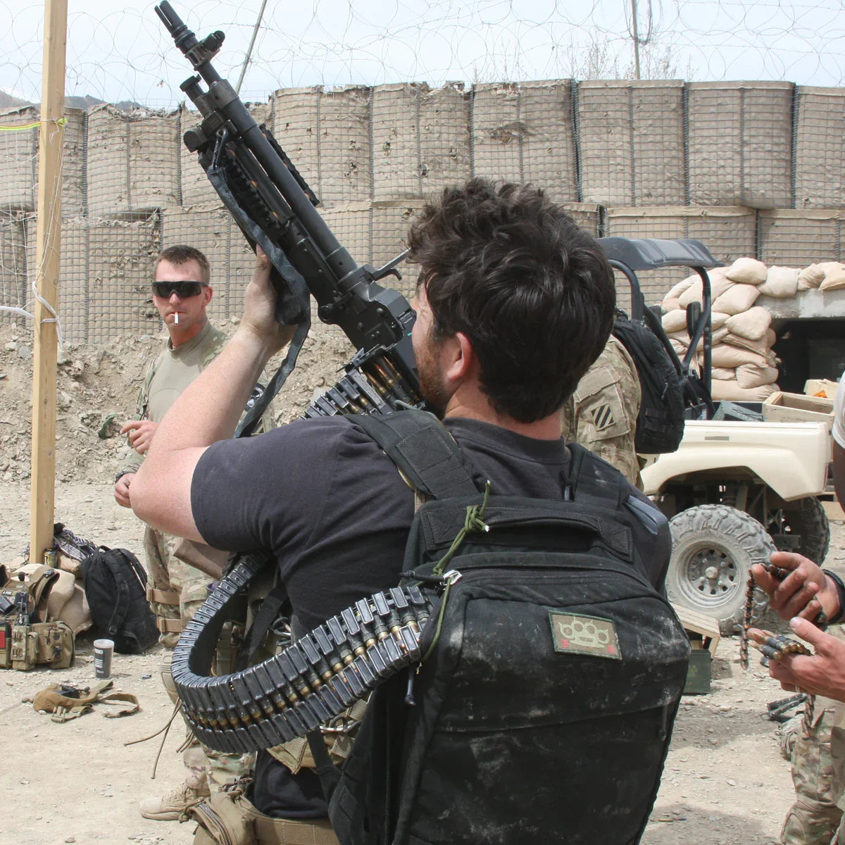 Man loading belt-fed machine gun with soldiers and military base background featuring sandbags and vehicle