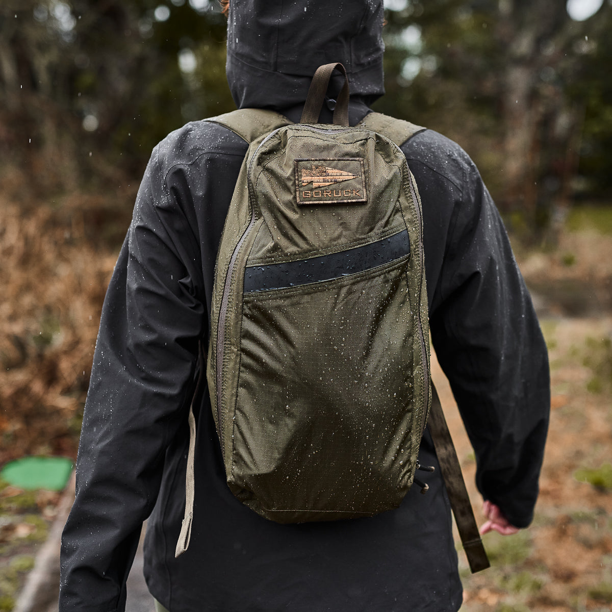 Person wearing a dark jacket and green backpack, walking outdoors in a rainy forest setting.
