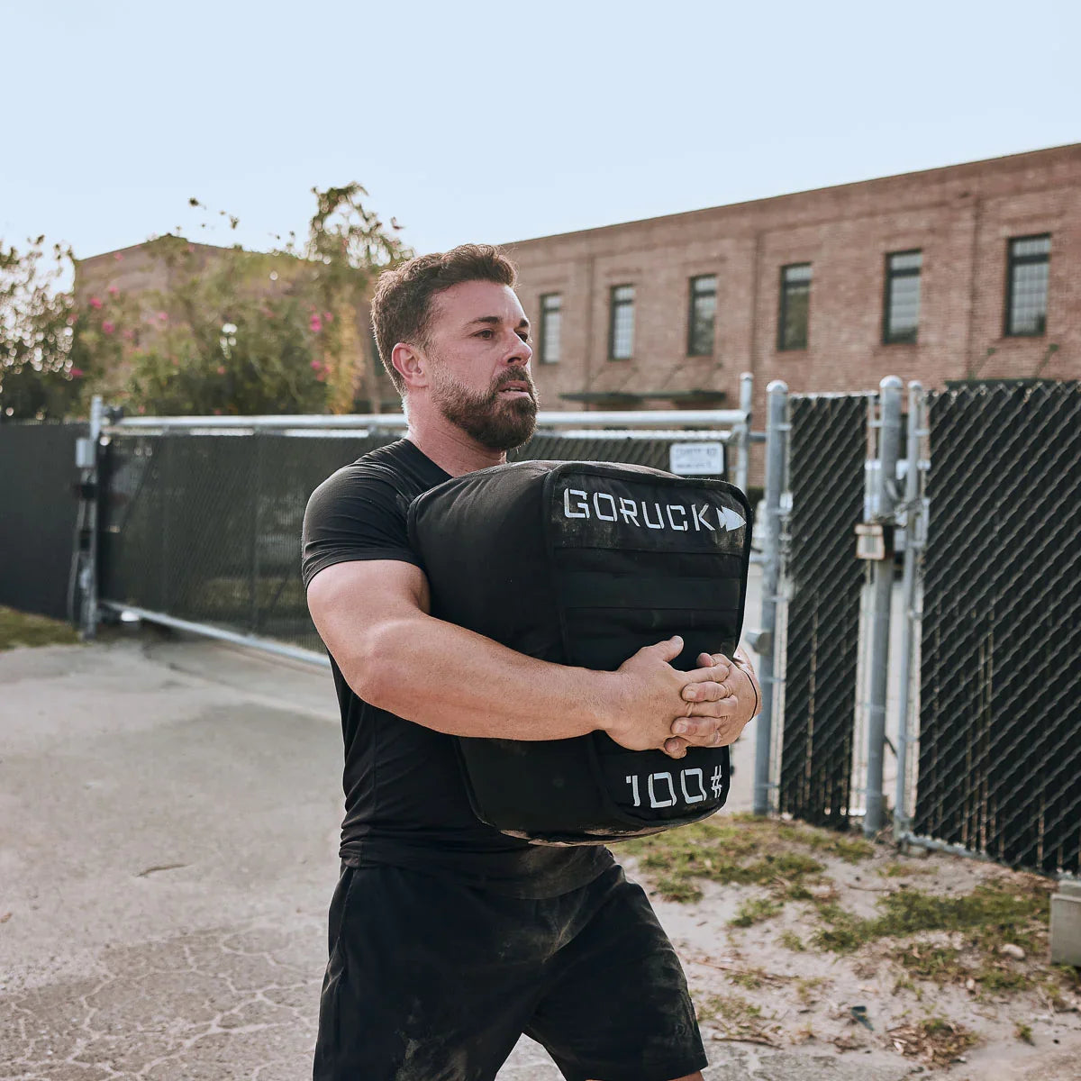 Fit man carrying a 100-pound GORUCK weighted sandbag outdoors near chain-link fence and brick building