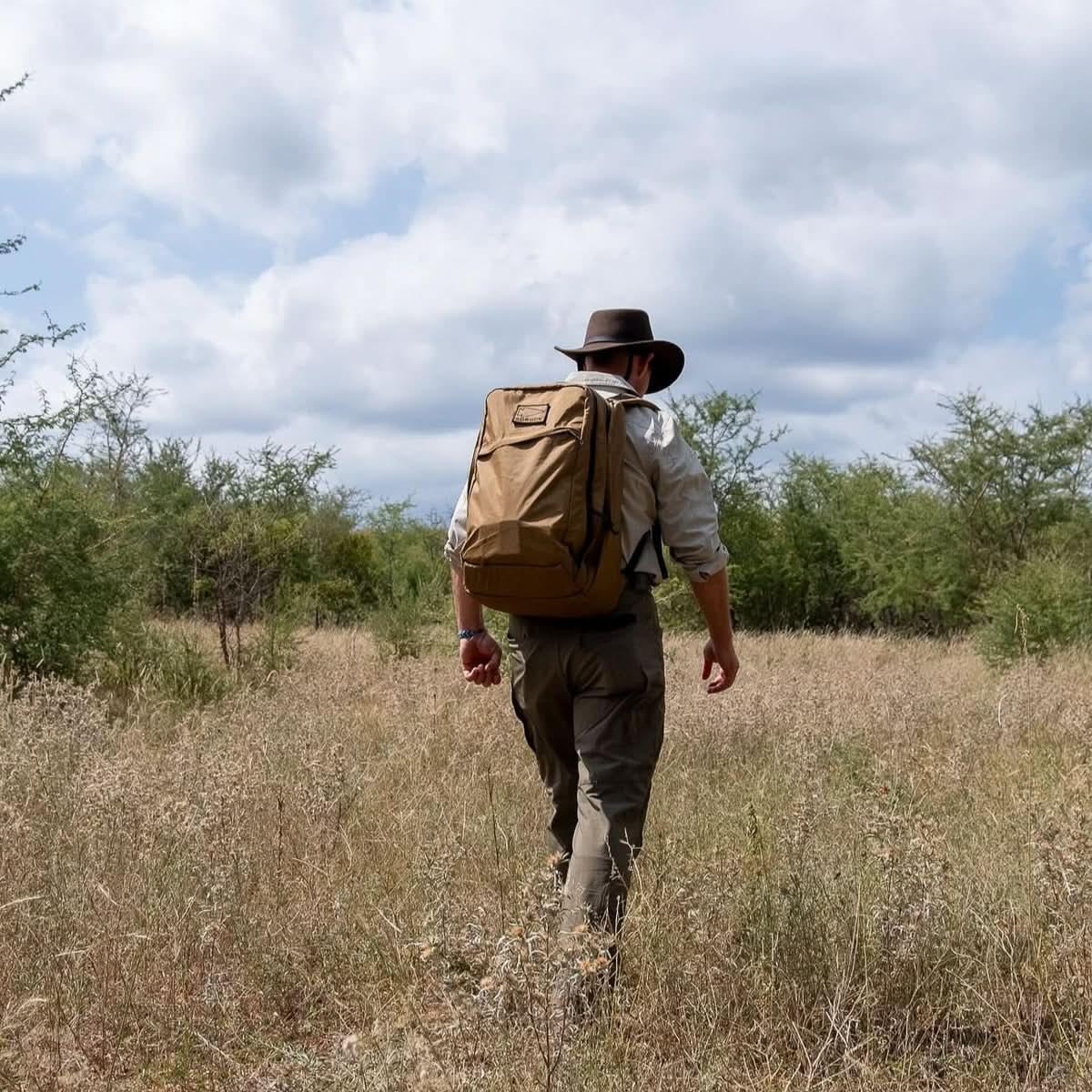 Person hiking through dry grassland wearing a brown backpack and wide-brim hat under cloudy sky