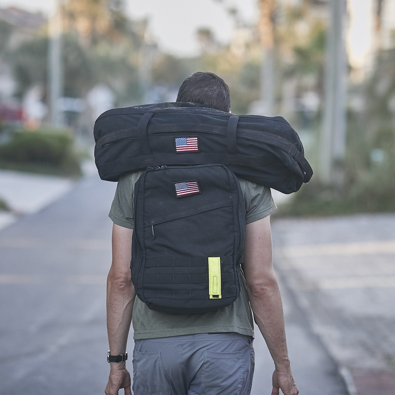 A person with a black backpack and a Sandbag Kit (w/ Filler Bag), both featuring US flag patches, walks down a street.