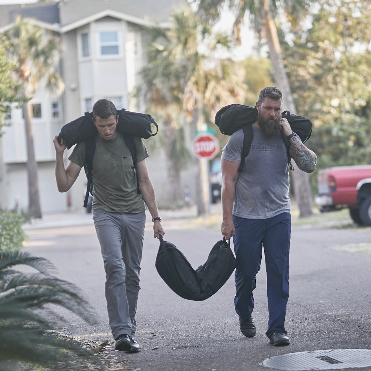 Two men carry Sandbag Kit (w/ Filler Bag) on their shoulders while walking down a residential street, pushing their limits with these durable training sandbags built to last.