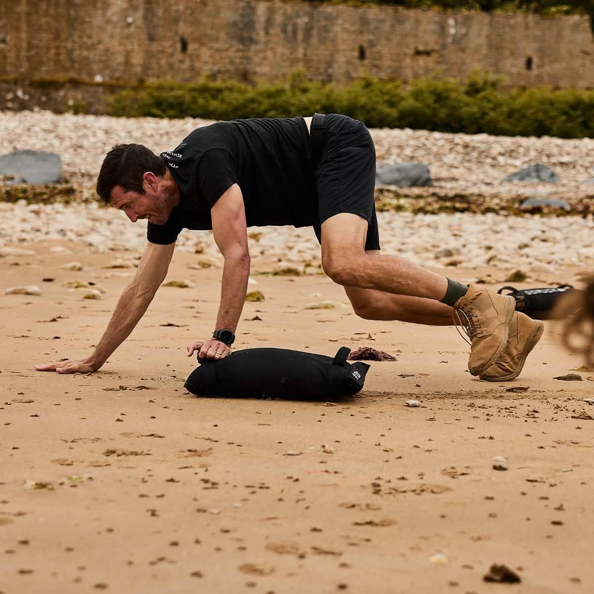 Man in black athletic gear doing high-intensity exercise with GORUCK sandbag on sandy beach