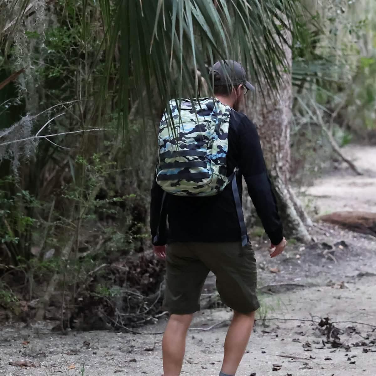 Man hiking in forest wearing black shirt, olive shorts, gray cap, and Everglades camouflage GORUCK backpack