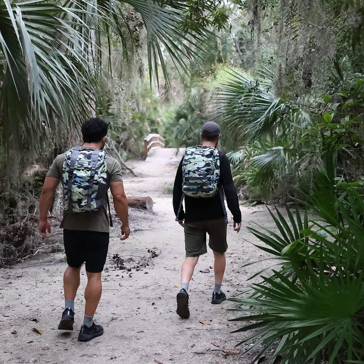 Two men rucking on a forest trail wearing Everglades camo backpacks in a lush, green outdoor setting