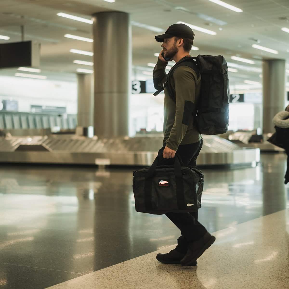 Man carrying GORUCK black rucksack and duffel bag in airport baggage claim area