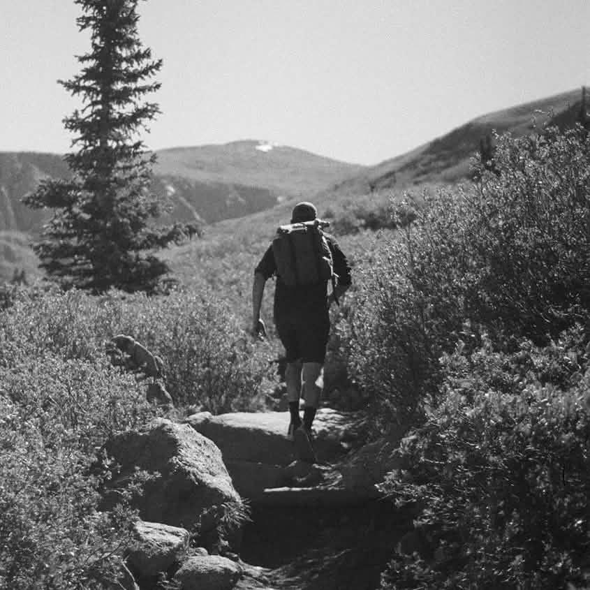 Hiker with GORUCK backpack walking on rugged mountain trail surrounded by bushes and rocks