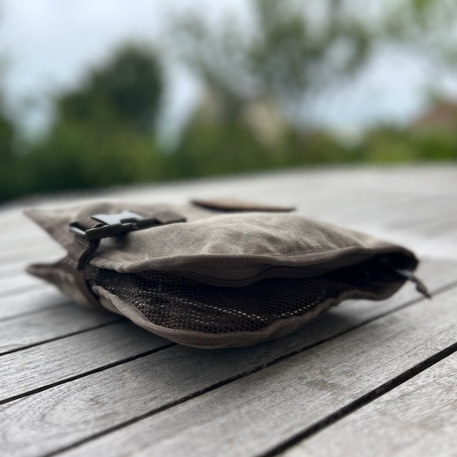 The Wire Dopp - Waxed Canvas, a compact brown pouch with a buckle, rests on a wooden table outdoors against blurred greenery—ideal for keeping your EDC organized.
