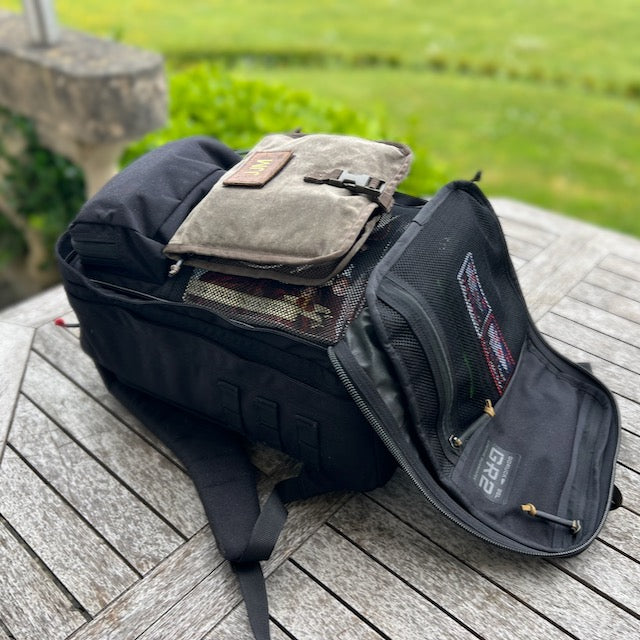 The Wire Dopp - Waxed Canvas sits atop a wooden outdoor table alongside a black backpack, offering a stylish and practical way to carry your EDC essentials.