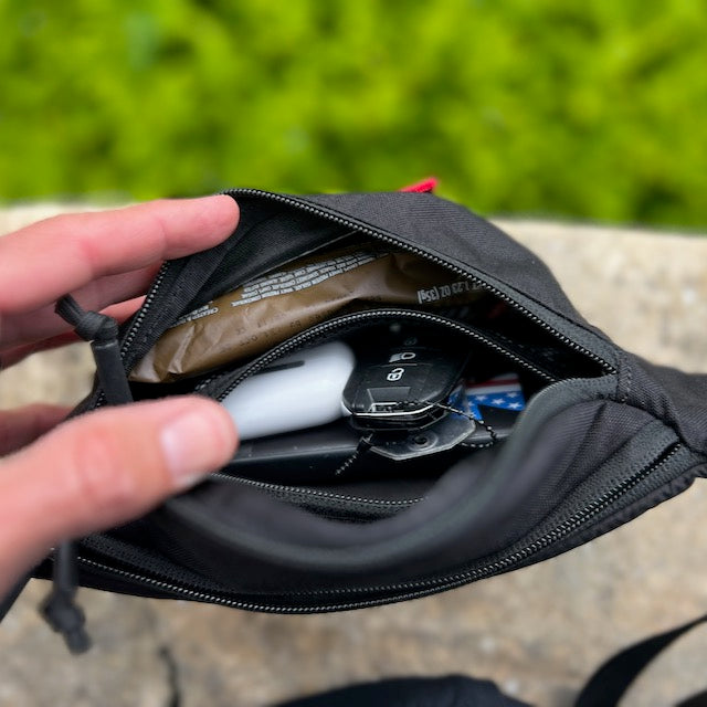 A hand opens the Tactical Fanny Pack - Cordura in black, revealing keys, a snack bar, AirPods, and other small essentials stored neatly inside.