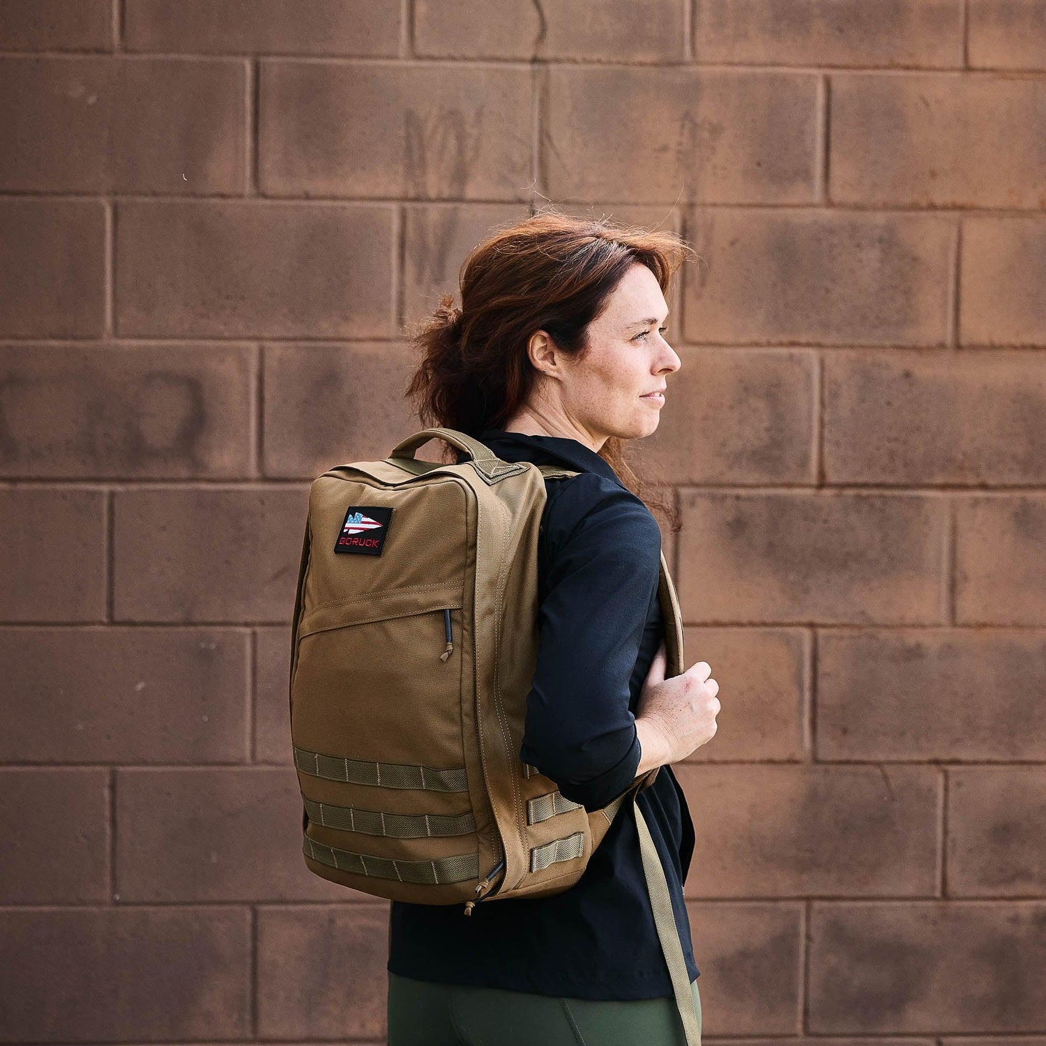 Woman wearing a tan GORUCK tactical backpack against a brown brick wall background