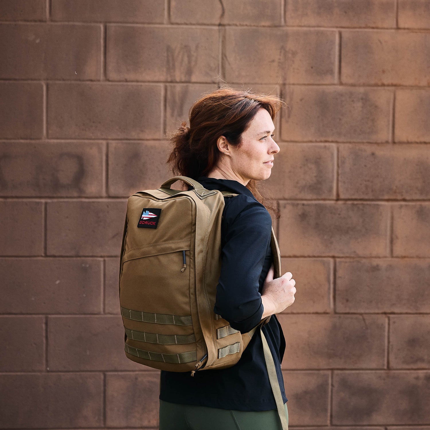 A woman wearing the GR1 USA - Cordura rucksack stands sideways before a brick wall, gazing into the distance.