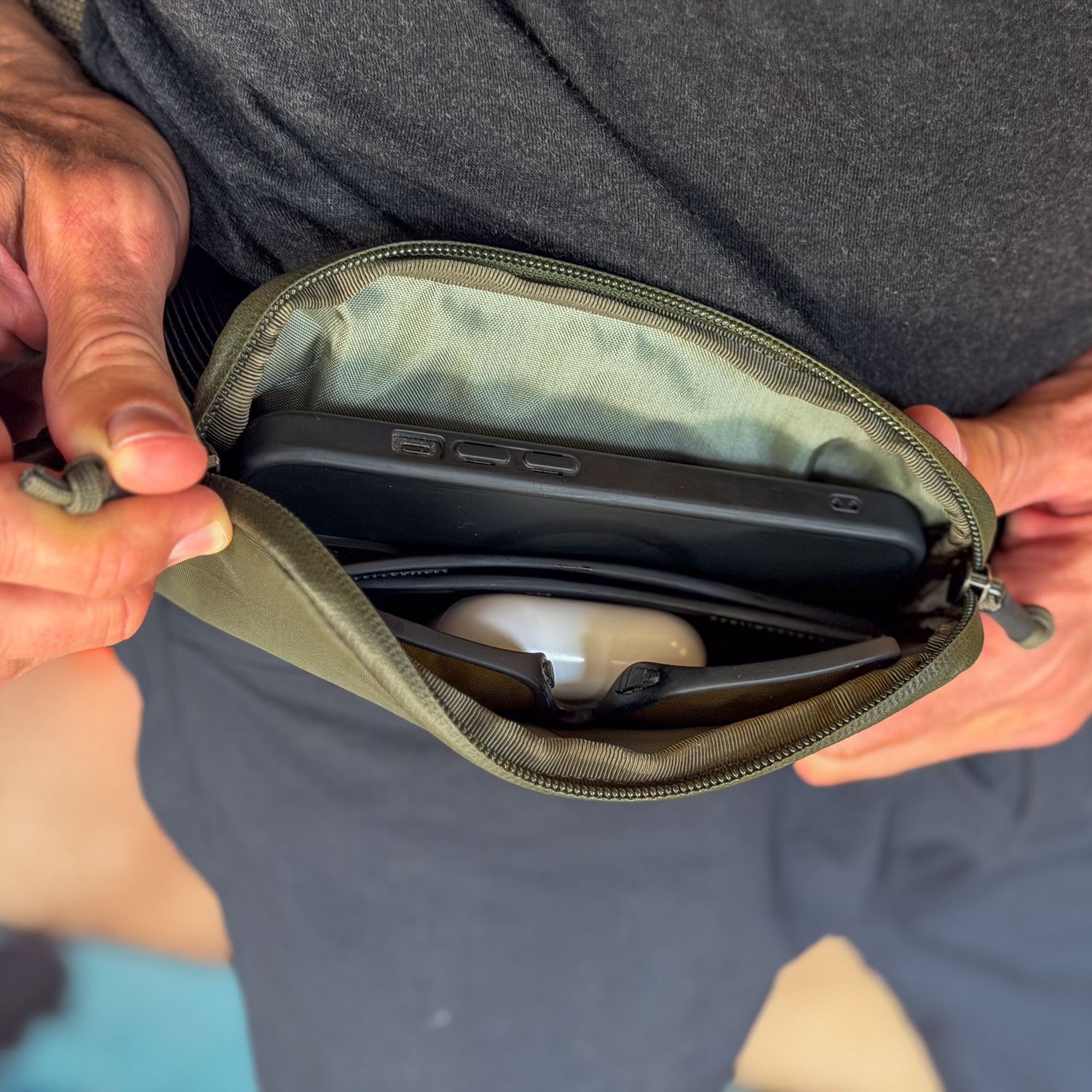 A person holding an open green Sternum / Belt Pouch - Waterproof X-PAC, revealing a smartphone and wireless earbuds—essential gear for those on the move.