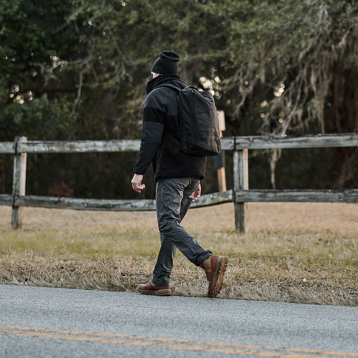 Wearing a beanie and dressed in dark clothing, a person walks along a wooden roadside fence. Their backpack suggests impending adventures, as they exude wanderlust and readiness in their Men’s Simple Pants - Midweight ToughDry®.