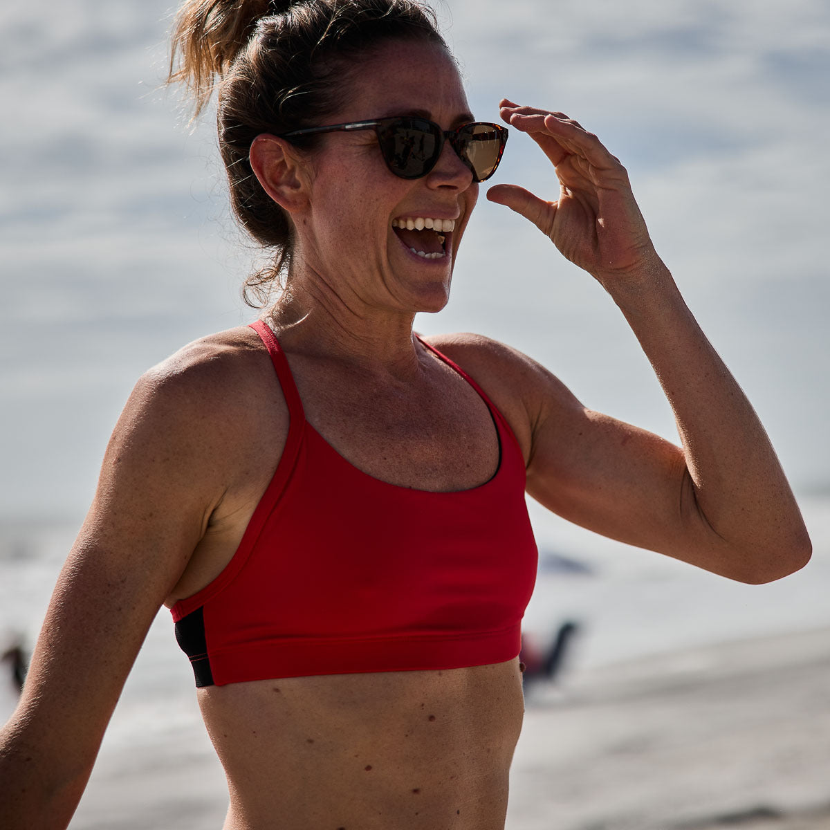 A woman wearing the Stealth Bra - ToughFlex and sunglasses smiles outdoors on a beach.