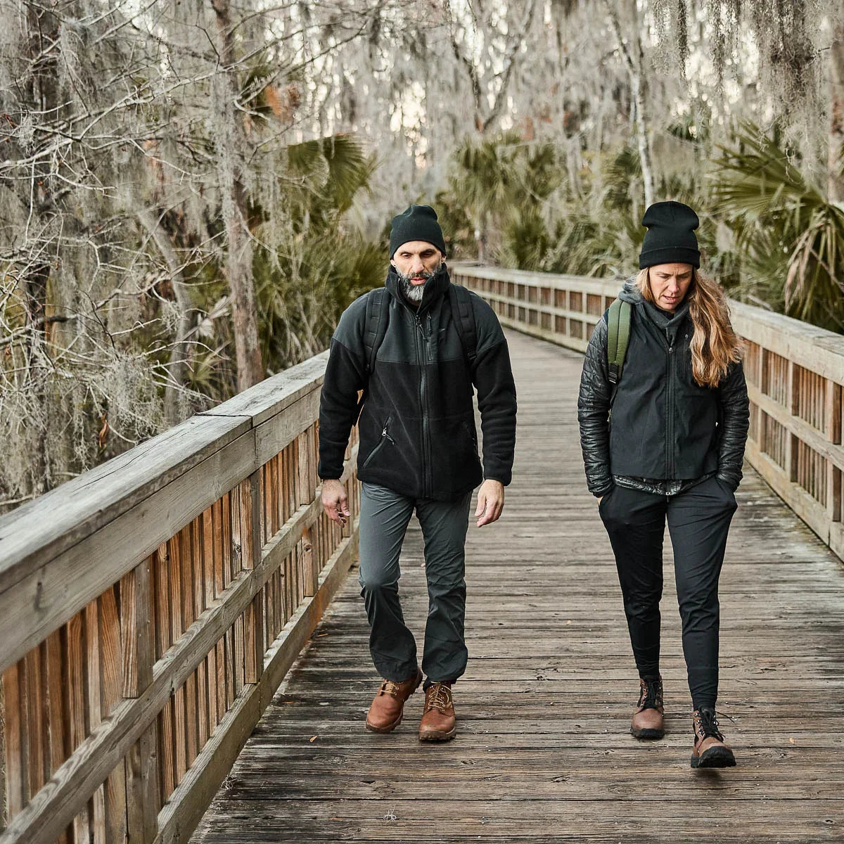 Two people wearing black jackets, backpacks, and boots walking on a wooden boardwalk through a mossy forest