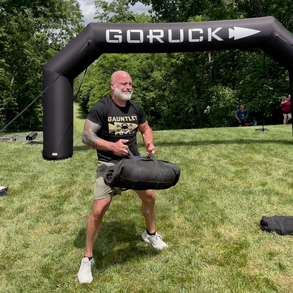 Wearing Merino Challenge Socks - Ankle, a man lifts a sandbag at an outdoor fitness event under a GORUCK arch on grass.