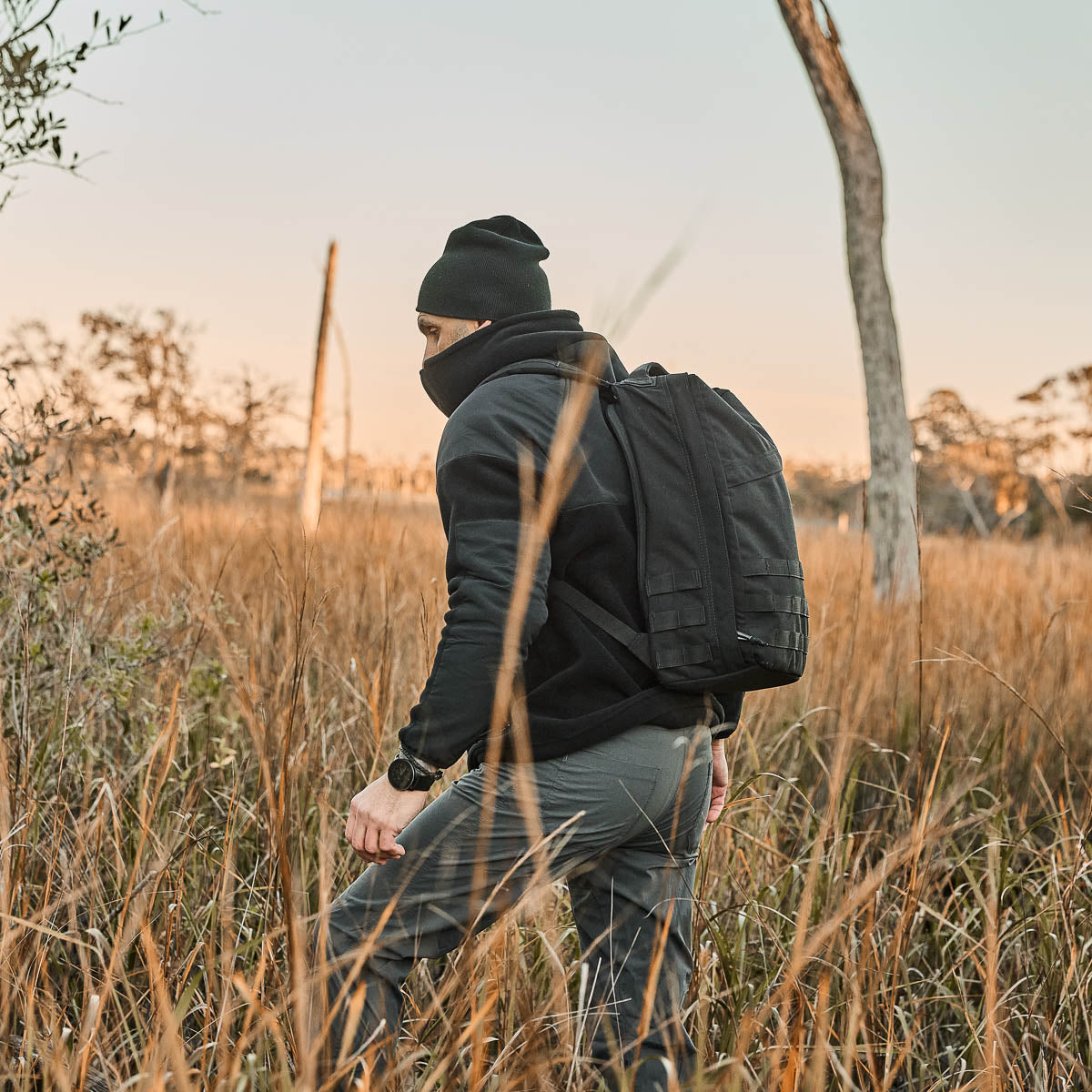 Wearing a black beanie and mask, someone in rugged Men’s Simple Pants - Midweight ToughDry® strolls through a grassy field at sunset with a backpack.