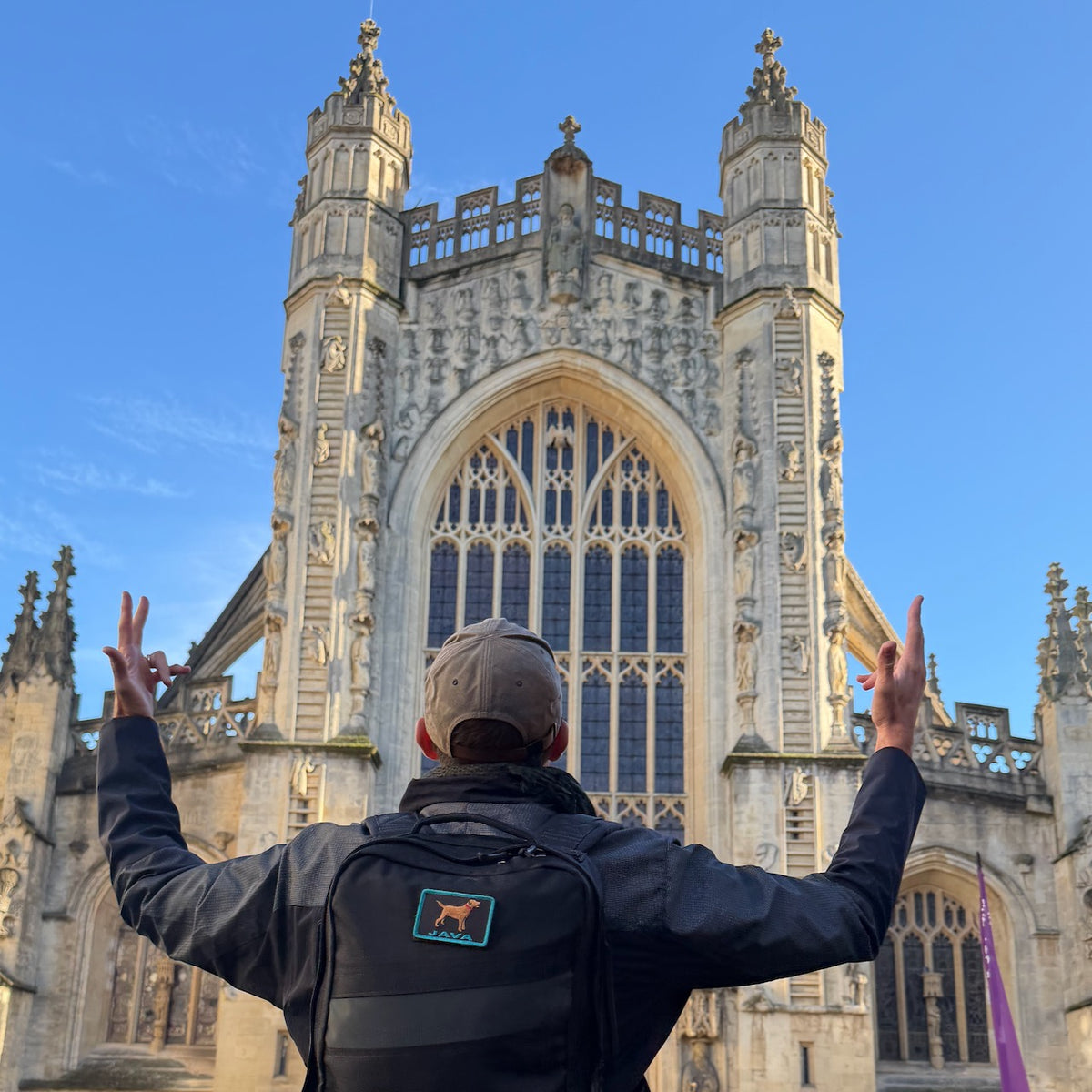 Wearing the Tac Hat - Heritage and a backpack, a person raises their hands skyward while facing a grand, ornate Gothic cathedral beneath a clear blue sky.