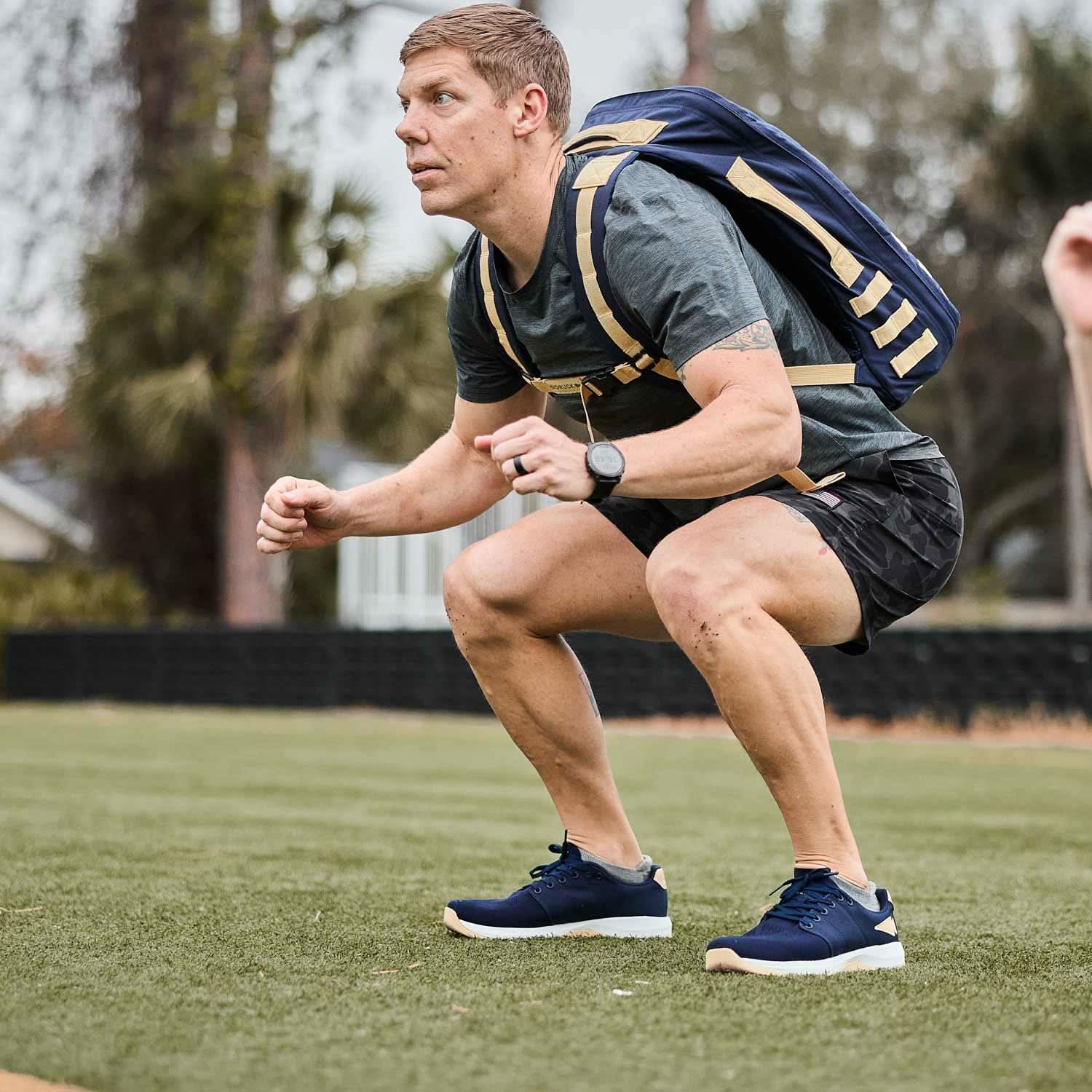 Man wearing GORUCK rucksack performing outdoor squat exercise on grass field