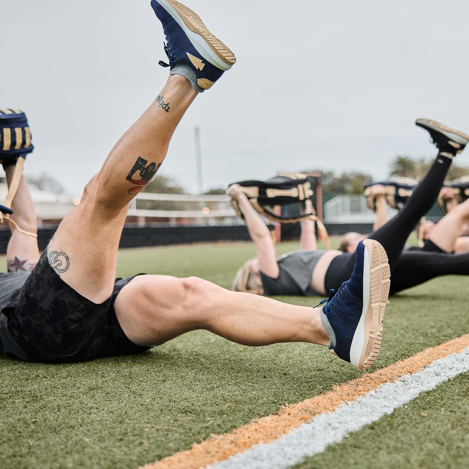 Group fitness session on grass field with people holding weighted bags and raising legs, wearing athletic shoes