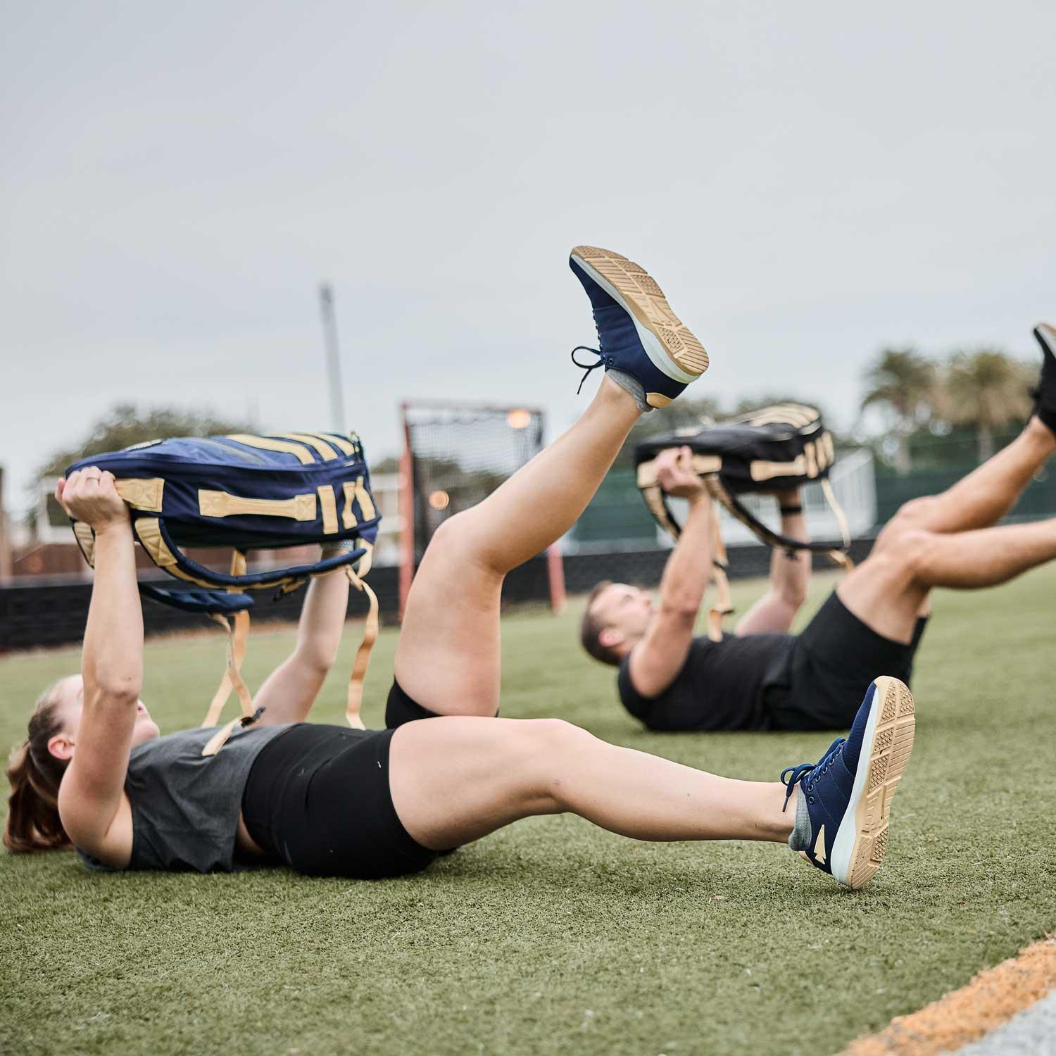 Two people doing leg raises on grass while holding blue and tan GORUCK backpacks in outdoor fitness training