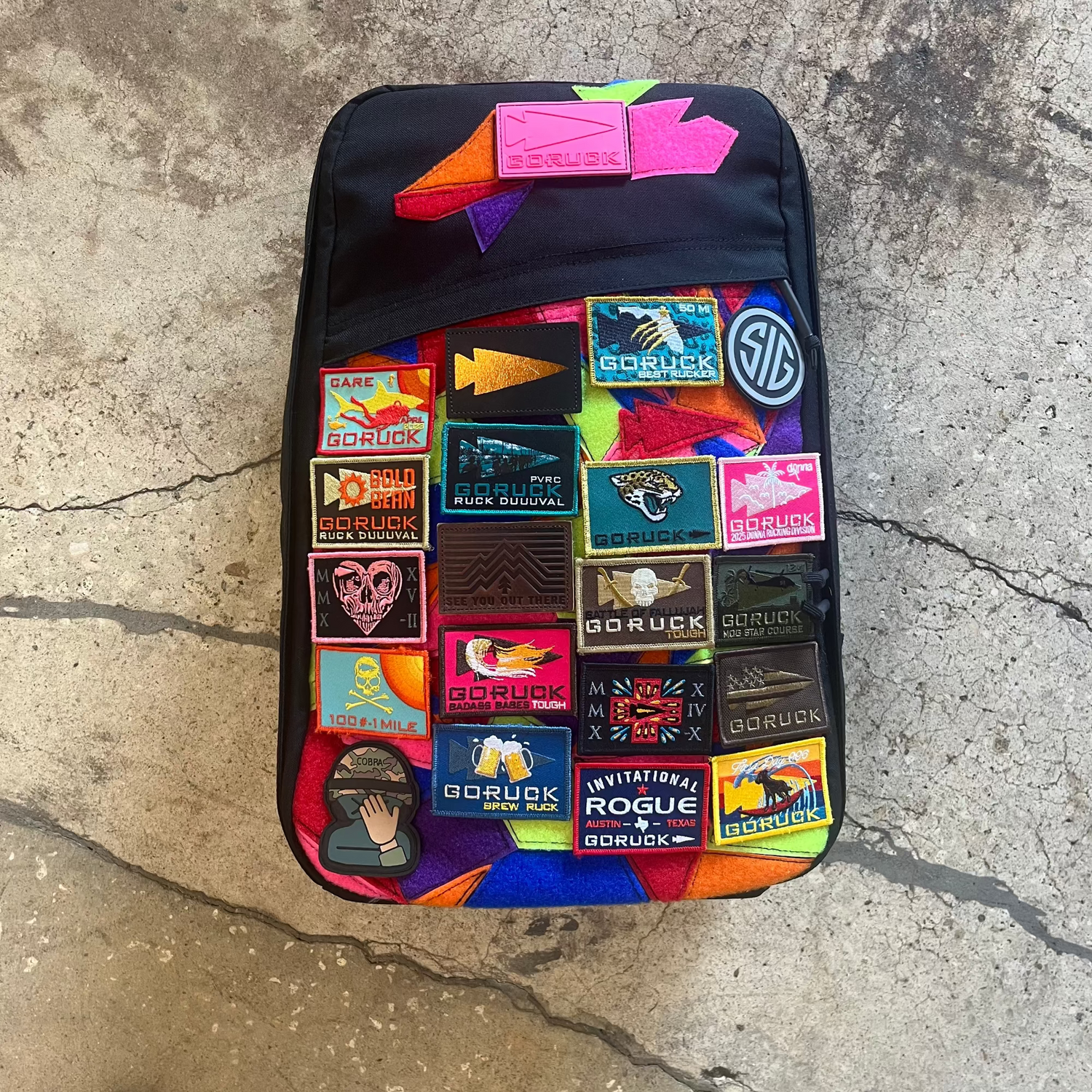 A black GR1 USA backpack made of ballistic nylon, adorned with colorful embroidered patches, rests on a cracked concrete floor.