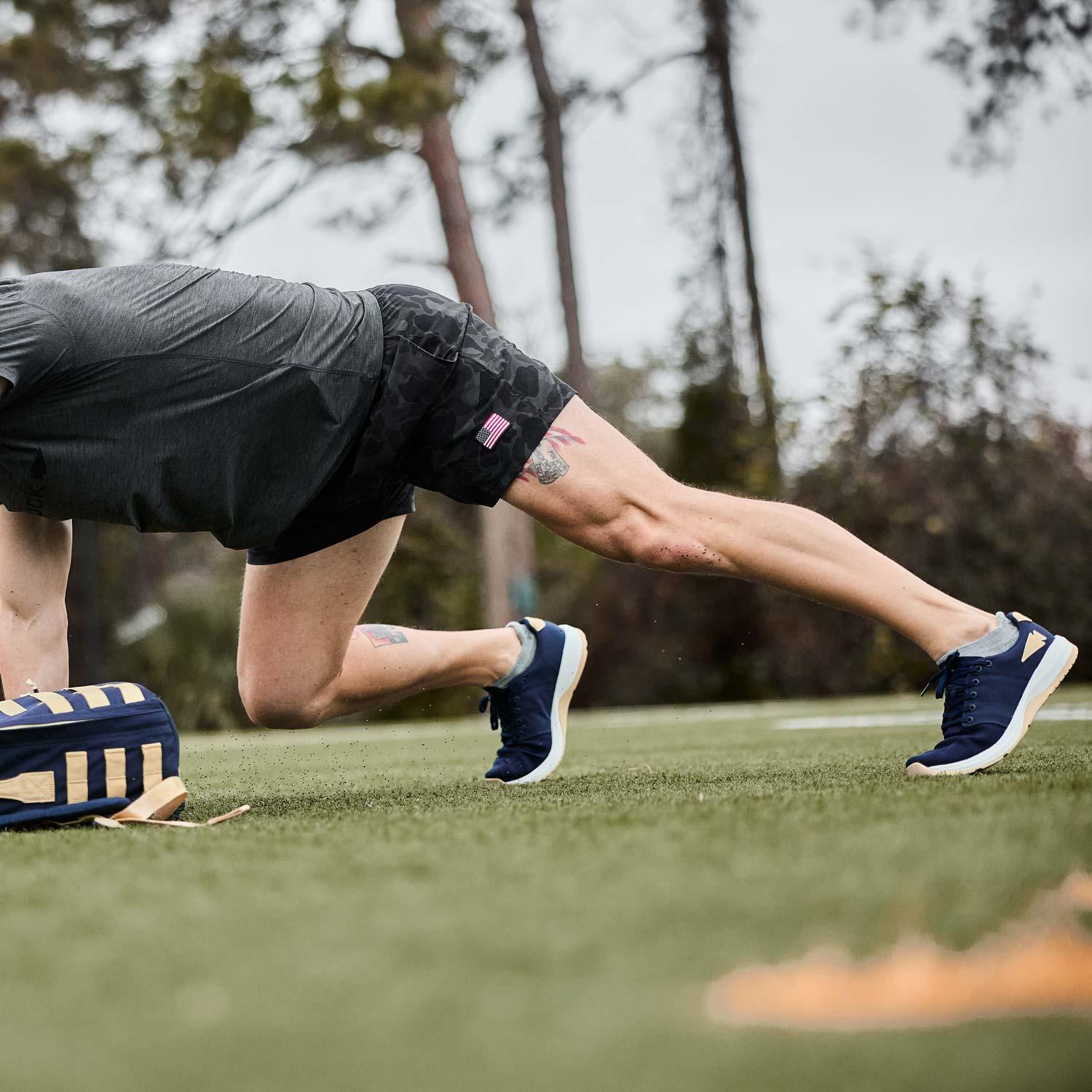 Athlete in black camo shorts and navy shoes performing a plank pose on grass near navy and tan rucking bag