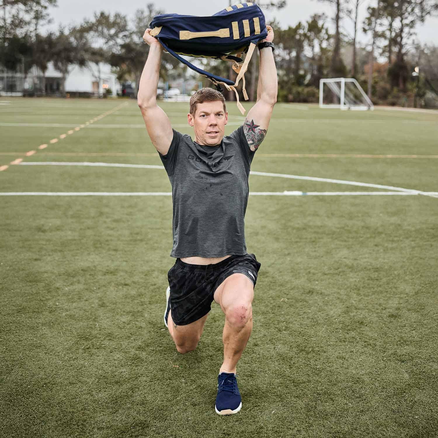 Athlete doing lunges with navy tactical rucksack overhead on green sports field with goal post