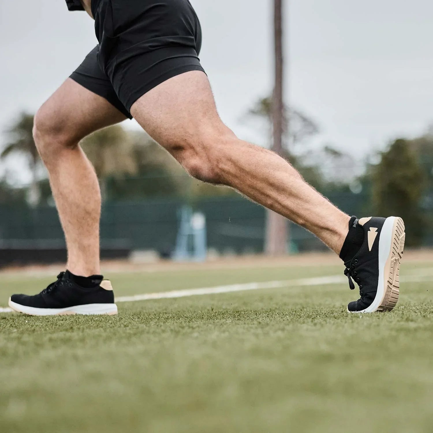 Close-up of a person's legs in black shorts and black athletic shoes stretching on green turf outdoors