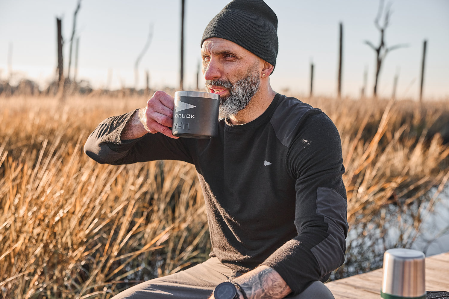 A bearded man in outdoor clothing sips from a mug while sitting in a grassy field, wearing the Men’s Commando Long Sleeve - Merino Wool shirt.