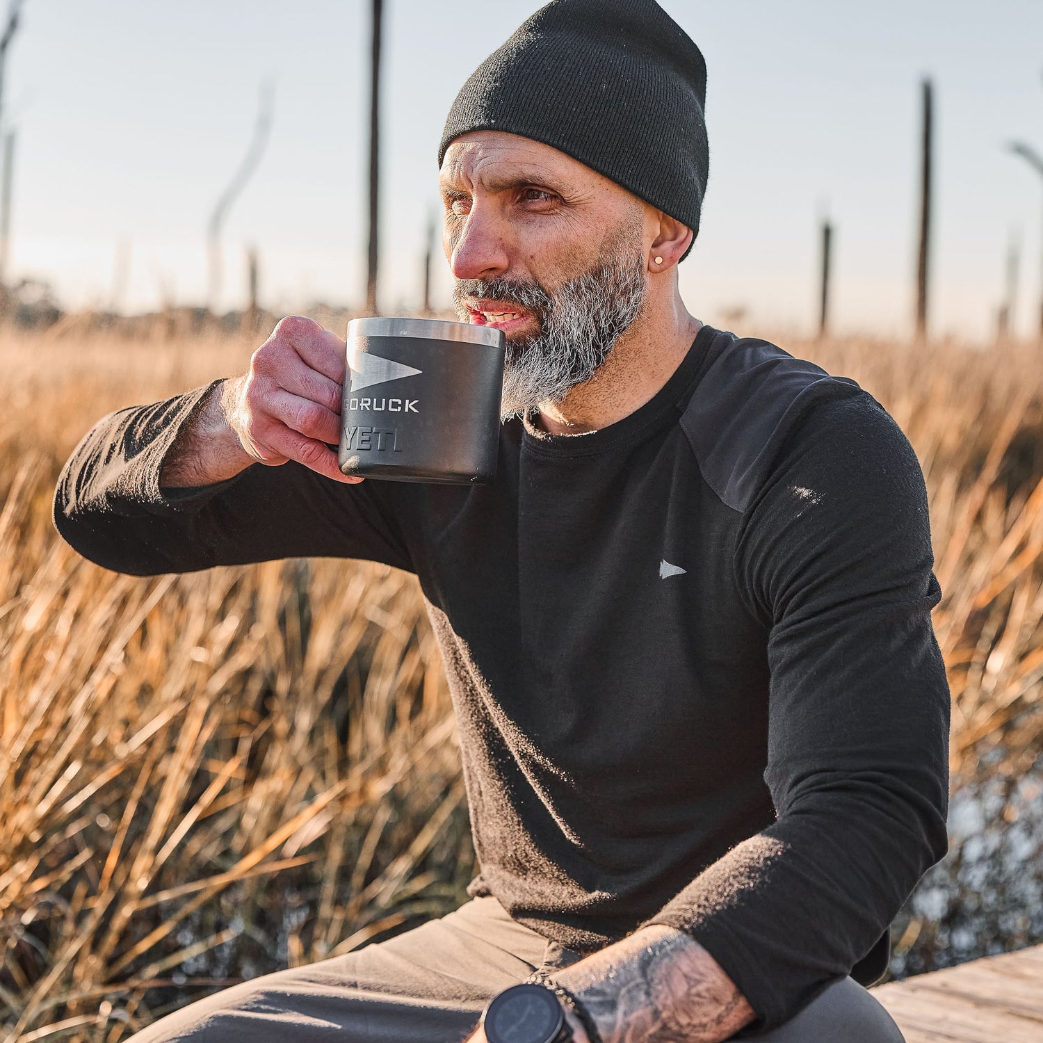 A man in a black beanie and the Men’s Commando Long Sleeve - Merino Wool sips from a mug while sitting outdoors by tall grass in sunlight.