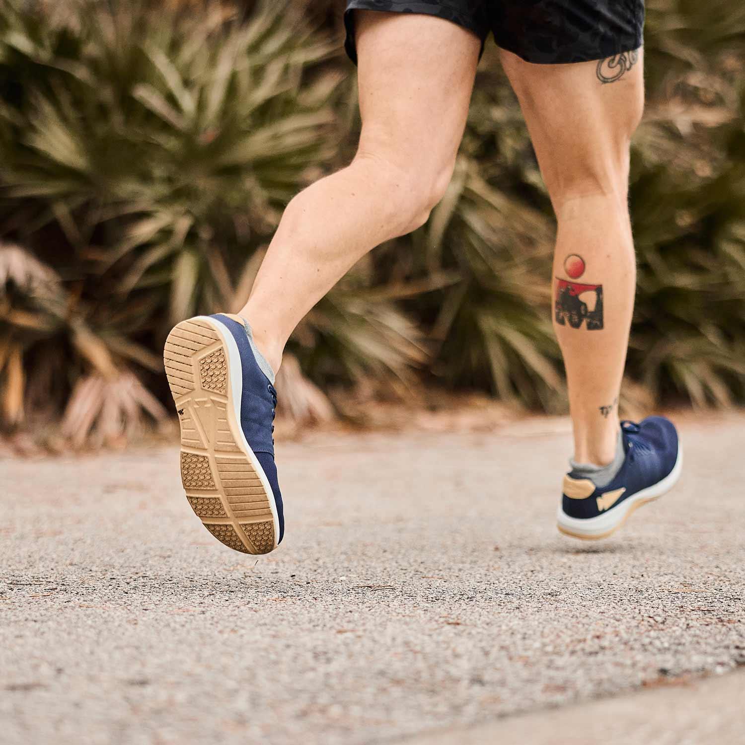 Close-up of a person running outdoors wearing blue athletic shoes and black shorts with tattoos on their legs