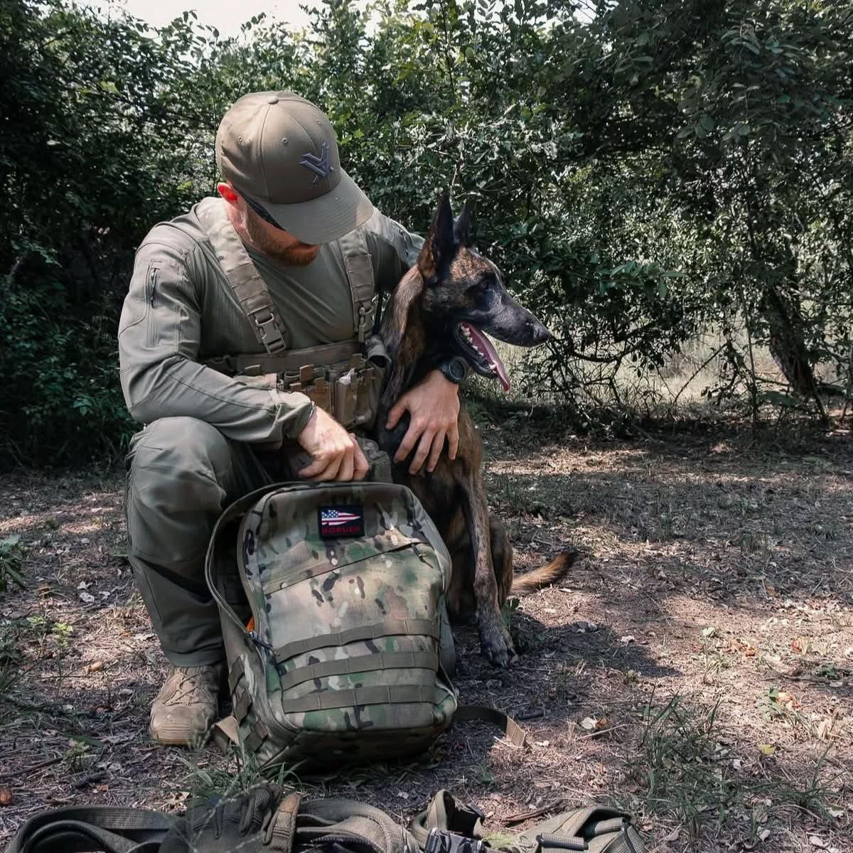 Man in tactical gear kneeling beside a military working dog outdoors with a camo GORUCK backpack