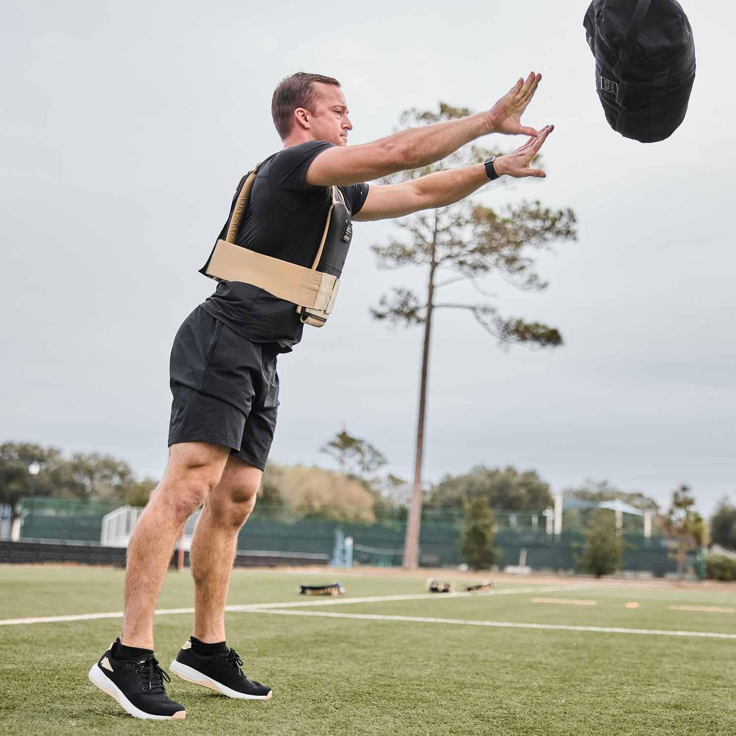 Man in rucking vest throwing a weighted sandbag outdoors on sports field with trees in background