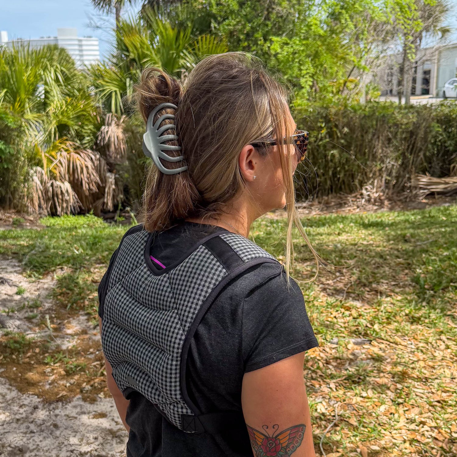 A woman with sunglasses and a hair clip wears a baby carrier and the Spy Ruck | Women's Weighted Vest, standing outdoors near greenery and buildings.