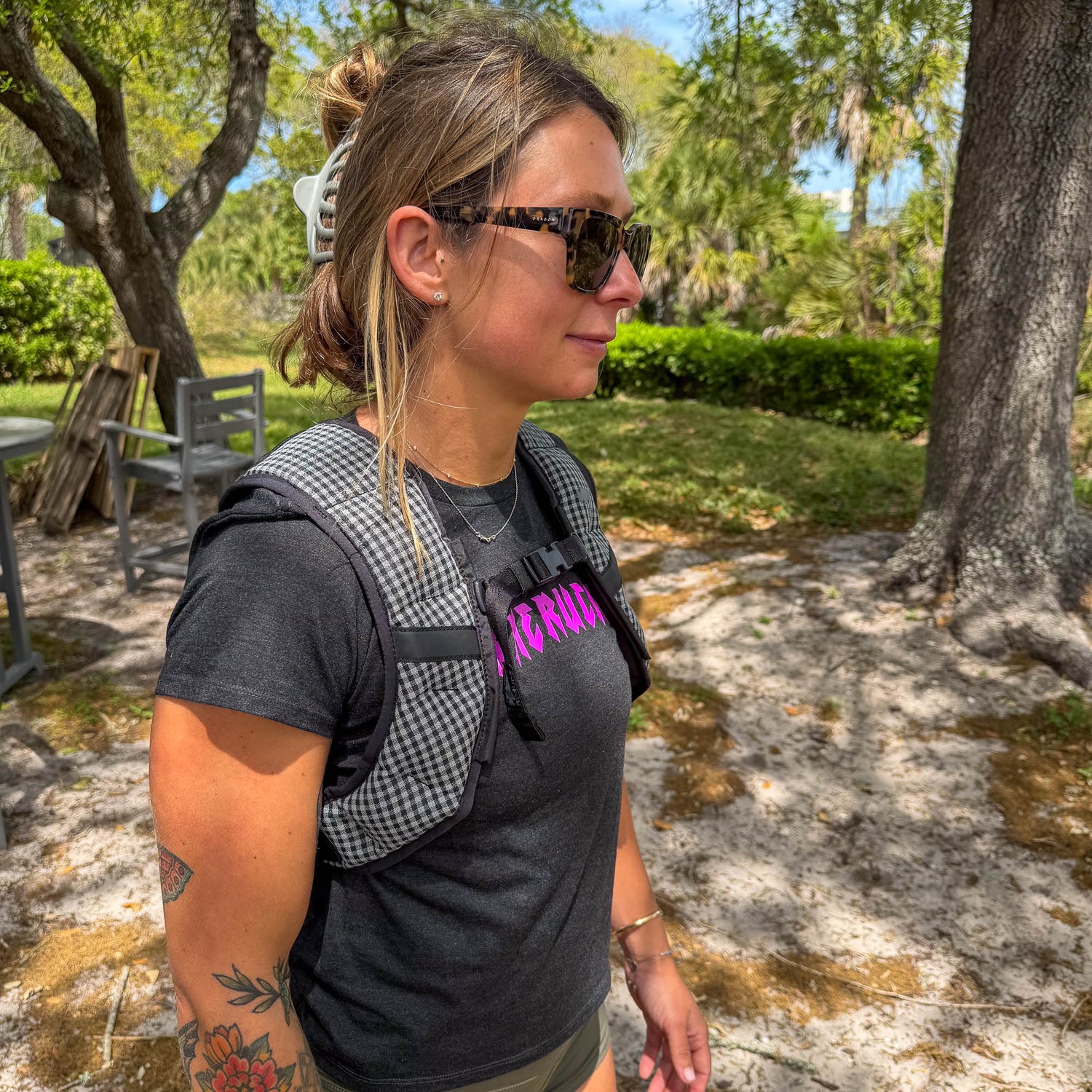 A woman wearing sunglasses and the Spy Ruck | Women's Weighted Vest stands outdoors in a sunny, wooded area surrounded by trees and greenery.