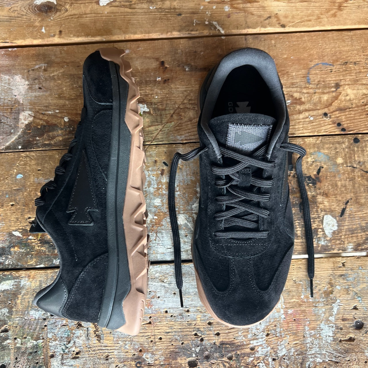 Mackall 45 Black + Gum sneakers with black suede uppers and tan rugged soles displayed on a worn, paint-splattered wooden surface.