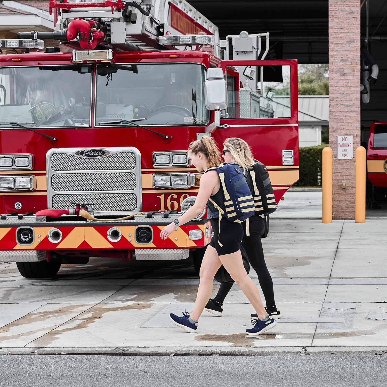 Two women rucking with GORUCK backpacks in front of a fire truck at a fire station