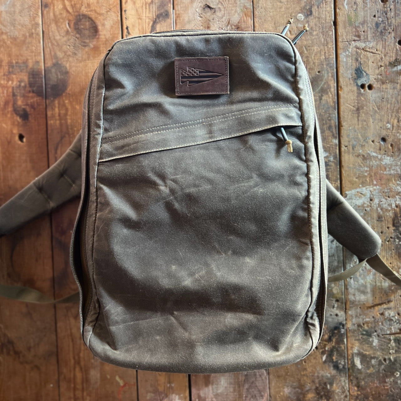 The GR1 USA - Heritage Waxed Canvas rucksack, made from brown waxed duck canvas and featuring a front pocket, rests on a rustic wooden surface.