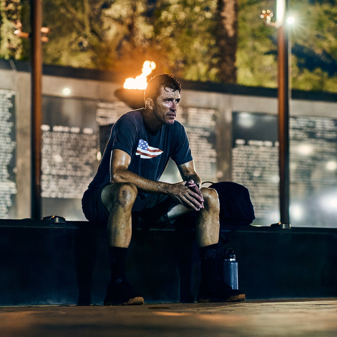 A sweaty man in athletic gear sits on a ledge at night, a flame and memorial wall behind him, wearing the GORUCK Flag Spearhead Tee - Unisex Tri-BlendX, its iconic spearhead design honoring Special Forces heritage.