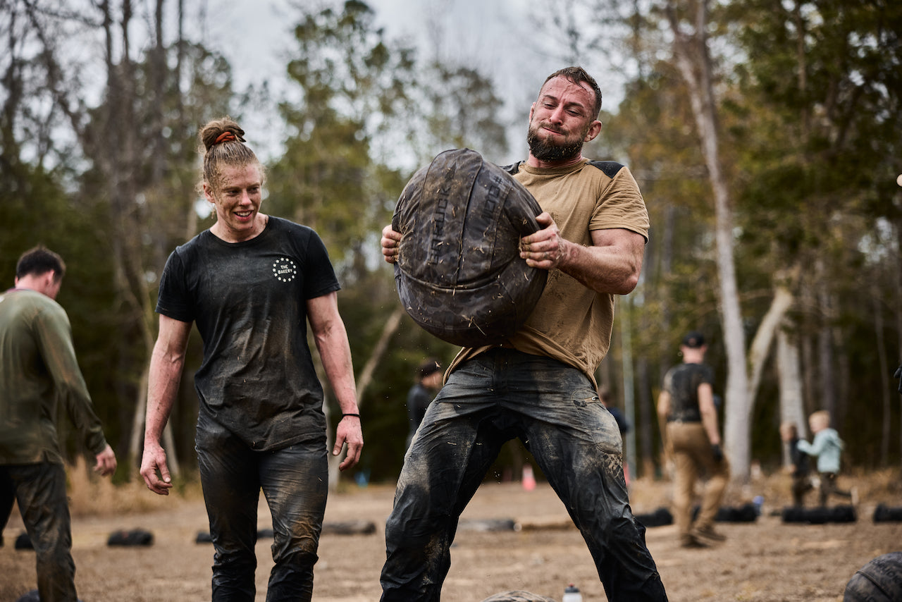 At Fort Bragg, two muddy competitors show off their grit as one lifts a heavy sandbag and the other smiles, embodying the challenge and spirit of BRAGG 2027.